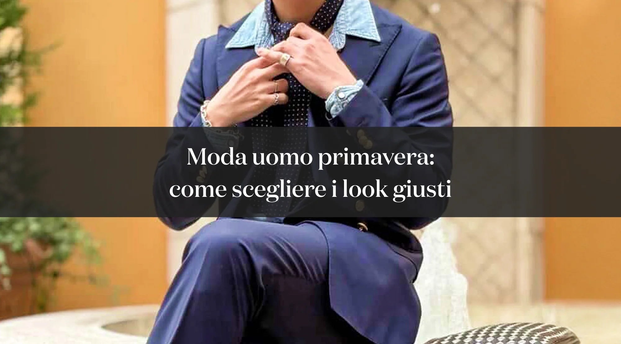 Uomo in abito blu seduto mentre sistema la cravatta, con sovrimpressione del testo “Moda uomo primavera: come scegliere i look giusti”